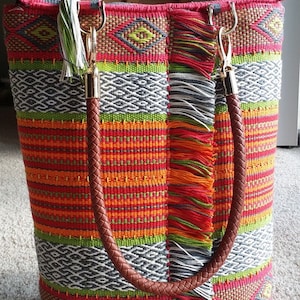 May include: A colorful woven tote bag with fringe details and leather handles. The bag has a geometric pattern in shades of red, orange, green, and gray.