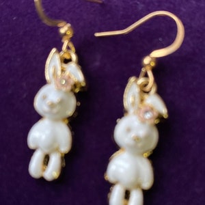 May include: A pair of gold dangle earrings featuring white bunny charms with pink flowers and a single rhinestone accent.