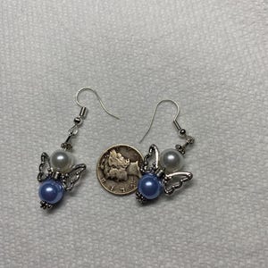 K&ouml;nnte beinhalten: Ein Paar silberne Engelsfl&uuml;gel-Ohrringe mit blauen und wei&szlig;en Perlenakzenten. Die Ohrringe werden neben einem US-amerikanischen Viertel f&uuml;r die Gr&ouml;&szlig;enreferenz gezeigt.