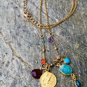 Puede incluir: Collar de cadena dorada con una variedad de piedras preciosas de colores, incluyendo rojo, naranja, azul, verde y morado. El collar presenta un colgante de moneda de oro con un dise&ntilde;o de una criatura m&iacute;tica.