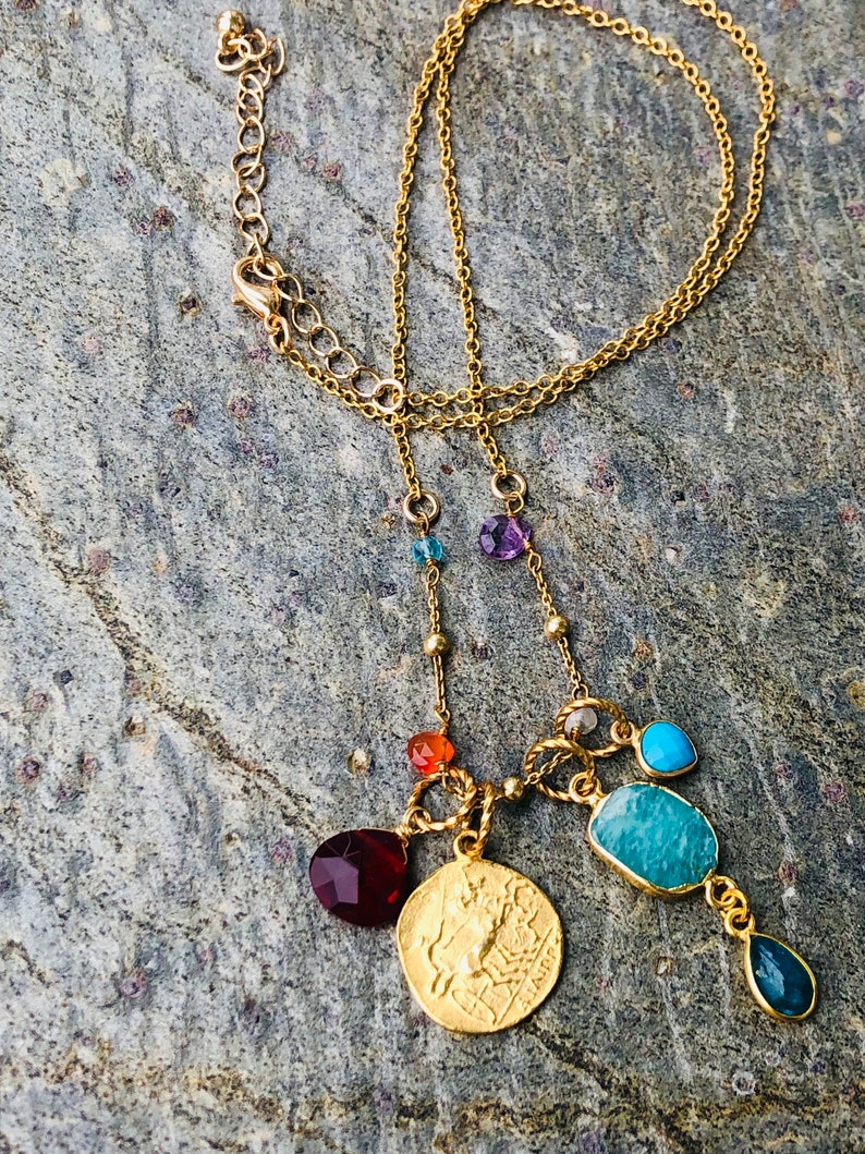 Puede incluir: Collar de cadena dorada con una variedad de piedras preciosas de colores, incluyendo rojo, naranja, azul, verde y morado. El collar presenta un colgante de moneda de oro con un dise&ntilde;o de una criatura m&iacute;tica.