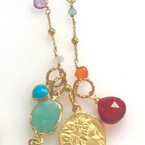 Puede incluir: Un collar de cadena dorada con m&uacute;ltiples dijes, que incluyen una moneda con una imagen de perfil, una l&aacute;grima turquesa, una piedra verde y piedras rojas y naranjas.