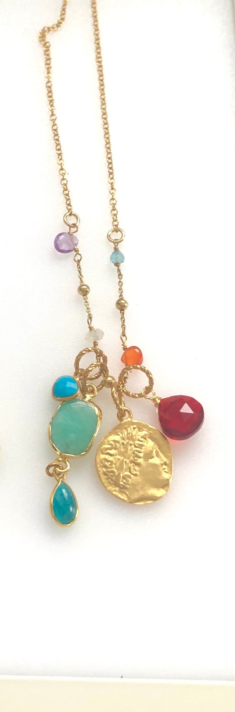 Puede incluir: Un collar de cadena dorada con m&uacute;ltiples dijes, que incluyen una moneda con una imagen de perfil, una l&aacute;grima turquesa, una piedra verde y piedras rojas y naranjas.