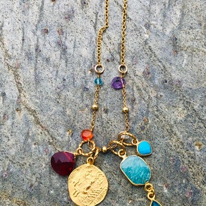 Puede incluir: Collar de cadena de oro con m&uacute;ltiples colgantes, incluyendo un colgante de moneda, una piedra turquesa y varias piedras preciosas m&aacute;s peque&ntilde;as en tonos de rojo, naranja, azul y morado.