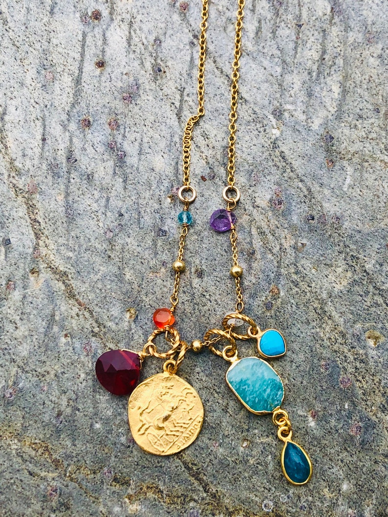 Puede incluir: Collar de cadena de oro con m&uacute;ltiples colgantes, incluyendo un colgante de moneda, una piedra turquesa y varias piedras preciosas m&aacute;s peque&ntilde;as en tonos de rojo, naranja, azul y morado.