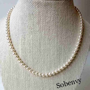May include: A white pearl necklace on a white fabric background. The necklace is made of small, round pearls.