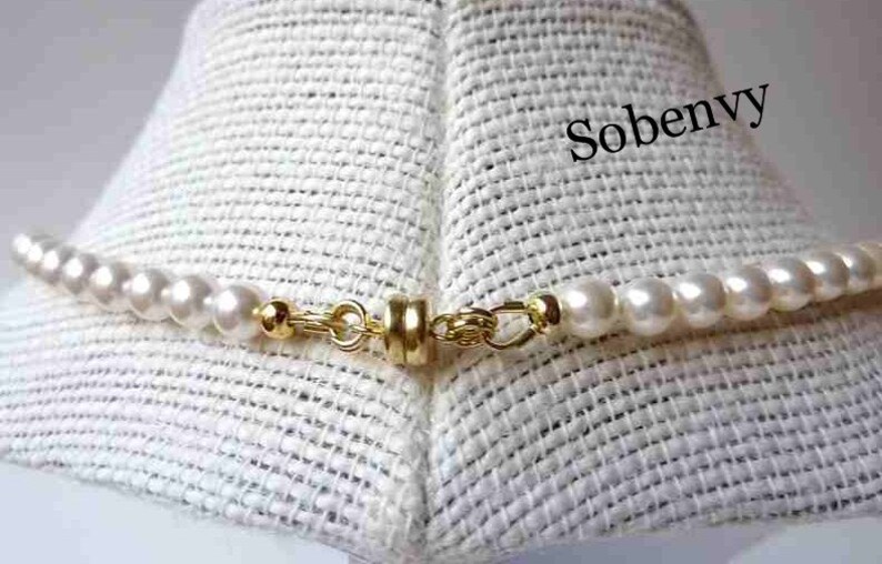 May include: A close-up of a white pearl necklace with a gold clasp. The necklace is on a white fabric background. The text "Sobenvy" is visible in the upper right corner of the image.