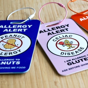 May include: A collection of colorful allergy alert tags. The tags are blue, pink, orange, and red, each with a different allergy warning. The tags have a metal ring for attachment and feature text and graphic symbols indicating specific food allergies, such as peanuts, gluten, and eggs.