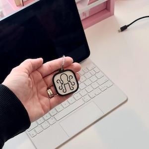 May include: A black and white octopus keychain with a white background and a black outline. The keychain is being held in a hand over a white keyboard.