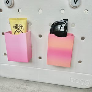 May include: A white tote bag with two pink and orange compartments. One compartment holds a tube of Sun Bum sunscreen, and the other holds black sunglasses. The bag has a perforated design and the word "bogg" is printed on the bottom.