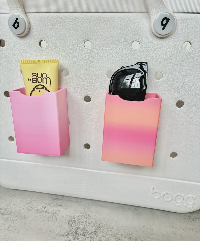 May include: A white tote bag with two pink and orange compartments. One compartment holds a tube of Sun Bum sunscreen, and the other holds black sunglasses. The bag has a perforated design and the word "bogg" is printed on the bottom.