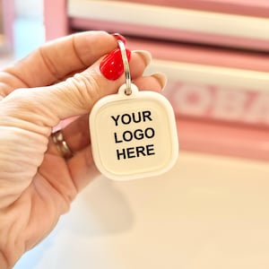 May include: White square keychain with a silver ring. The keychain has the text "YOUR LOGO HERE" printed in black.