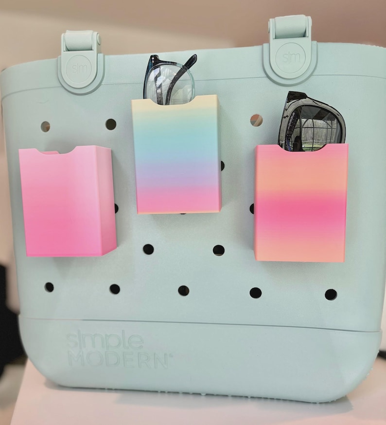May include: A pale blue tote bag with three colourful, rectangular pockets attached. Each pocket has a gradient of colours, and two pairs of spectacles are stored in two of the pockets. The bag has the words "simple MODERN" printed on the front.