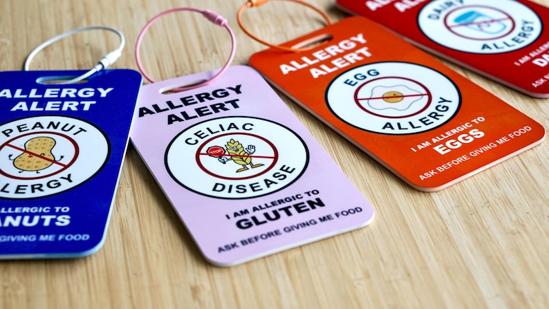 May include: A collection of colorful allergy alert tags. The tags are blue, pink, orange, and red, each with a different allergy warning. The tags have a metal ring for attachment and feature text and graphic symbols indicating specific food allergies, such as peanuts, gluten, and eggs.