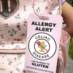 May include: A pink allergy alert tag with the text "ALLERGY ALERT" and "CELIAC DISEASE". The tag includes the message "I AM ALLERGIC TO GLUTEN" and a warning to "ASK BEFORE GIVING ME FOOD". The tag is attached to a pink floral backpack.