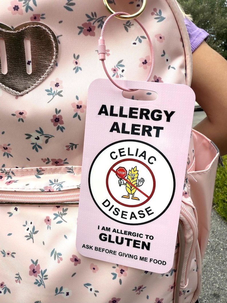 May include: A pink allergy alert tag with the text "ALLERGY ALERT" and "CELIAC DISEASE". The tag includes the message "I AM ALLERGIC TO GLUTEN" and a warning to "ASK BEFORE GIVING ME FOOD". The tag is attached to a pink floral backpack.