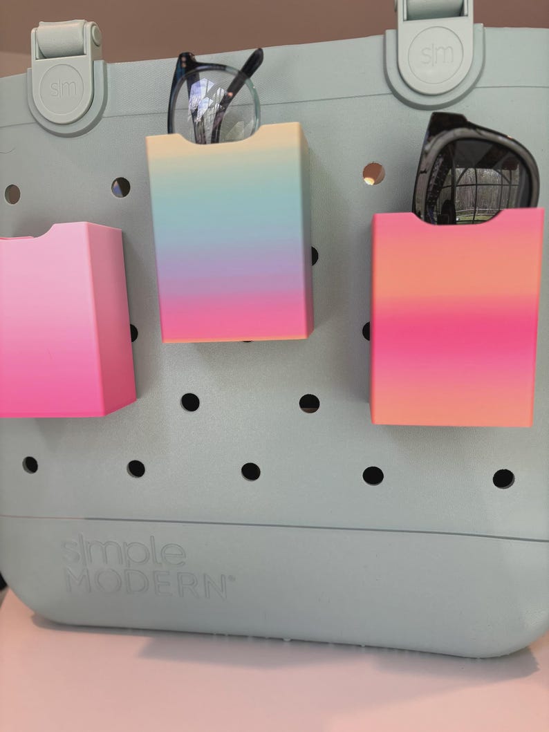 May include: A pale blue tote bag with three gradient-coloured storage pockets. One pocket is pink, and the other two are a mix of pink, orange, blue, and yellow. Eyeglasses and sunglasses are stored in the pockets. The bag has the words "Simple Modern" printed on it.