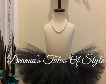 May include: A black tulle tutu skirt on a mannequin. The skirt is full and fluffy, and it has a satin waistband. The text "Deanna's Tutus Of Style" is written in white cursive on the skirt.