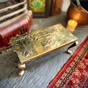 Puede incluir: Un reposapiés rectangular de latón con detalles florales en relieve. El reposapiés tiene cuatro patas cortas y redondeadas. La superficie superior presenta rosas y hojas en relieve. Se coloca sobre una superficie de piedra gris junto a una alfombra roja estampada.