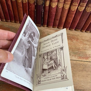 May include: A burgundy leather-bound book with the title "Adam Bede" by George Eliot. The book is open to a page with an illustration of a woman fainting and a man sitting in a chair reading a book. The text "Look there! she's fainting" is visible on the page.