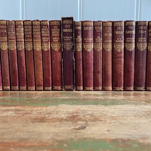 May include: A collection of 24 vintage hardback books with maroon covers and gold lettering. The books are arranged in a row on a wooden surface.