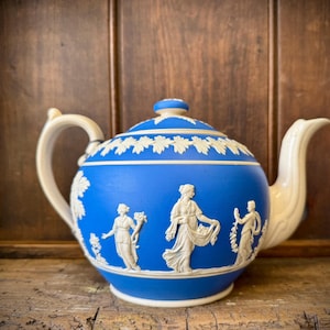 Könnte beinhalten: Blaue Keramik-Teekanne mit weißen Relief-Figuren und floralen Akzenten. Die Teekanne hat einen gebogenen Ausguss und Henkel sowie einen Deckel mit kleinem Knauf. Die Figuren sind im klassischen Stil gehalten.