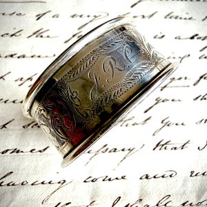 May include: A silver napkin ring with engraved floral and leaf patterns. The initials "J.R.C" are etched into the metal. The ring is set against a backdrop of handwritten script.