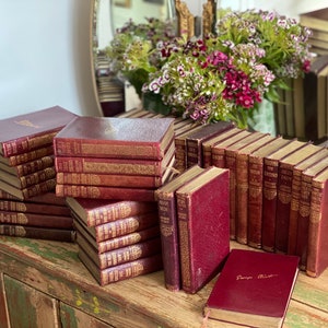 May include: A collection of vintage, burgundy-colored books with gold lettering and decorative accents. The books are stacked on a wooden table.