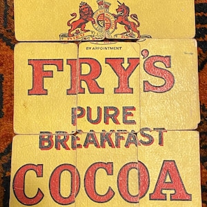 Peut inclure: Une boîte vintage de cacao pur pour le petit-déjeuner Fry's. La boîte présente un blason royal avec les mots "By Appointment". Le texte "Fry's Pure Breakfast Cocoa" est en lettres rouges sur un fond jaune.