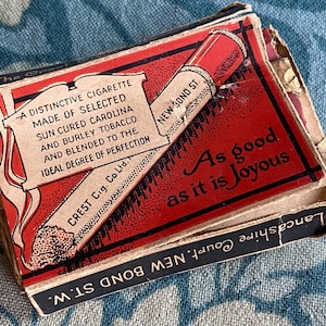 May include: Vintage Crest Cigarettes box with a red and white design. The box features text that reads "A distinctive cigarette made of selected sun cured Carolina and Burley tobacco." The text "As good as it is joyous" is also visible.