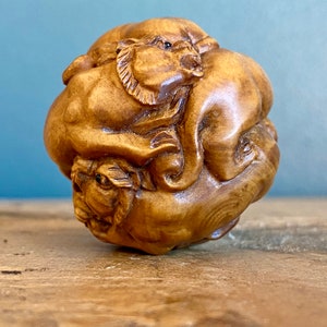 Puede incluir: Una escultura de madera de una bola con cuatro caras de animales talladas en ella. Los animales son un león, un tigre, un oso y un dragón. La escultura está hecha de madera marrón claro y tiene un acabado liso.