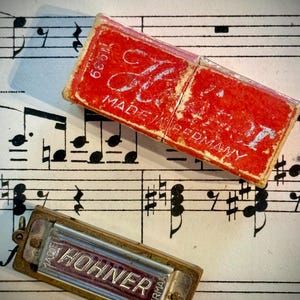 May include: A vintage Hohner harmonica with a metallic body and a separate red cardboard box featuring the text "MADE IN GERMANY" and "1939". The harmonica has a silver-toned plate with the brand name "HOHNER" embossed in a dark red centre.