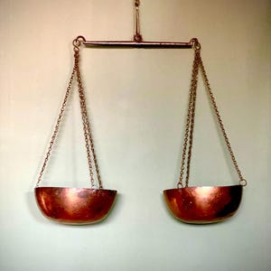 Op de afbeelding: Een vintage koperen balansweegschaal met twee hangende kommen. De weegschaal is gemaakt van metaal en heeft een eenvoudig ontwerp.