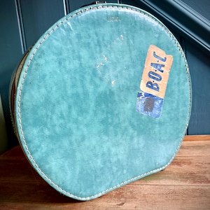 Può includere: Una valigia da viaggio vintage, rotonda, di colore turchese, con un bordo cucito bianco. La valigia ha un aspetto usurato e un adesivo "BOAC" sbiadito. È appoggiata su una superficie di legno invecchiato.