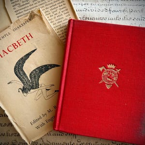 May include: Two antique books are shown. One is titled "Macbeth" with a winged figure illustration. The other is a red book with a gold emblem. The books rest on a backdrop of aged paper with handwritten text.