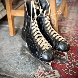 May include: A pair of vintage black ice skates with cream laces. The skates are on a stone surface with a red patterned rug. The skates have metal blades and are near a wooden stool. The skates are a classic design.