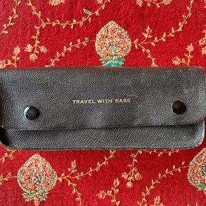 Puede incluir: Una funda de viaje de cuero negro con letras doradas que dicen "TRAVEL WITH EASE". La funda tiene dos broches metálicos en la parte delantera.