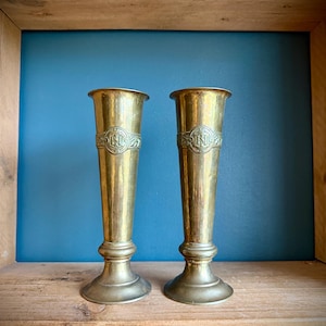 May include: Two antique brass vases with a flared top and a decorative band. The vases have a pedestal base and are displayed against a blue background. Each vase is approximately 25 cm tall.