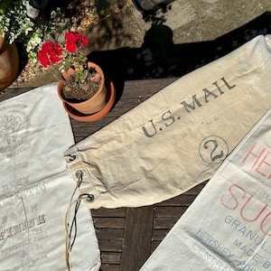 May include: A collection of vintage canvas sacks and a small terracotta pot with red flowers. One sack is a U.S. Mail bag, another reads "HERSHEY SUGAR", and a third has text in a decorative font.
