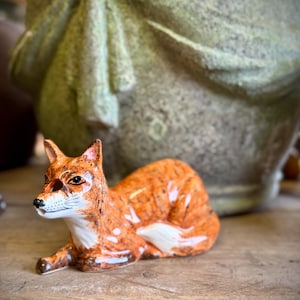 Puede incluir: Una figurita de cerámica de un zorro acostado. El zorro está pintado en tonos naranja y marrón con detalles blancos. La figurita está apoyada sobre una superficie de madera.