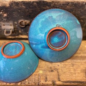 Op de afbeelding: Twee teal keramische kommen met een bruine rand. De kommen staan op een houten oppervlak.