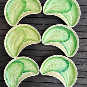 Puede incluir: Seis platos de ensalada de cerámica con un diseño de hoja de lechuga. Los platos tienen forma de media luna, con un borde color crema y un patrón de hoja de lechuga verde. Los platos están colocados sobre una superficie de madera oscura.