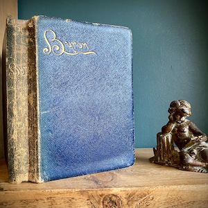 May include: A vintage blue hardcover book with gold lettering that reads "Byron" on the spine. The book is sitting on a wooden shelf next to a small bronze statue of a person.