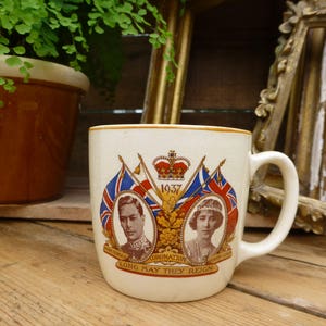 Może przedstawiać: Kremowy ceramiczny kubek z portretami królewskimi, flagami i koroną, z napisem "1937" i "Long May They Reign". Kubek ma zakrzywiony uchwyt i złoty brzeg, upamiętniający koronację.