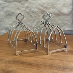 May include: Two silver-toned metal toast racks featuring four arched slots each. The racks have a rectangular base with four small feet and a central handle at the top shaped like a stylized quatrefoil. The metal has a polished, reflective finish.