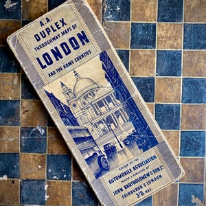 May include: A vintage, tan-coloured map book with a blue cover. The cover features a black and white illustration of St. Paul's Cathedral in London. The title of the book is "A.A. Duplex Throughway Maps of London and the Home Counties." The book is published by John Bartholomew & Son Ltd. and is priced at 3/6 net.