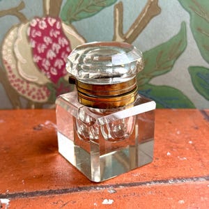 Op de afbeelding: Een inktpot van helder glas met een vierkante voet en een gefacetteerde, ronde glazen stop. De stop wordt vastgehouden door een metalen, messingkleurige kraag met een getextureerde, geribbelde rand. De inktpot heeft een hol, rond reservoir.