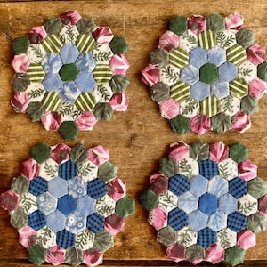 Pode incluir: Quatro  apoios de copo de tecido feitos à mão com um design hexagonal. Cada apoio de copo apresenta um centro azul e verde rodeado por um padrão de tecido rosa, verde e branco.