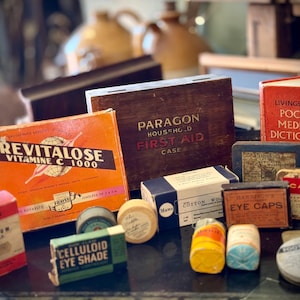 May include: A selection of antique medical items, featuring a box of "Revitalose Vitamin C 1000", a wooden "Paragon Household First Aid Case", and a red "Livingstone's Pocket Medical Dictionary". Also present are small boxes and tins with various labels.
