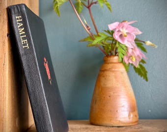 Op de afbeelding: Een zwart boek met de titel "HAMLET" in gouden letters, rustend op een houten plank. Een klein rood embleem is zichtbaar op de kaft. Daarnaast staat een kleine, bruine vaas met roze bloemen en groene bladeren.